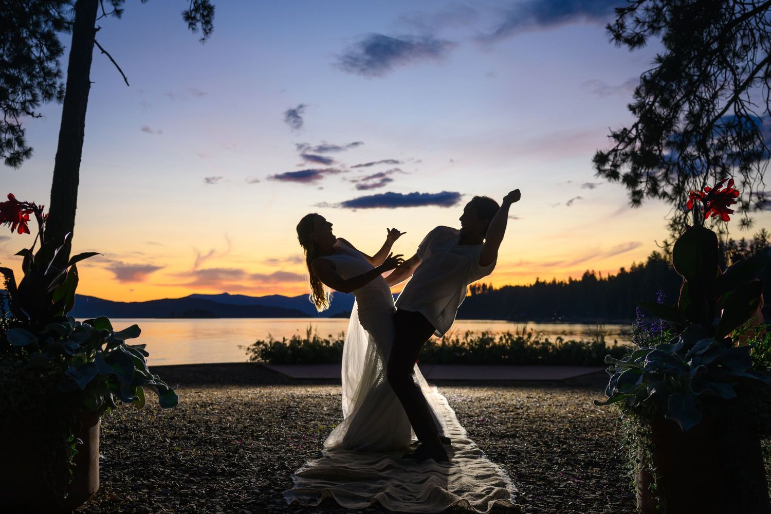 Silhouette dancing at sunset by a serene lake with mountains and trees in the background.