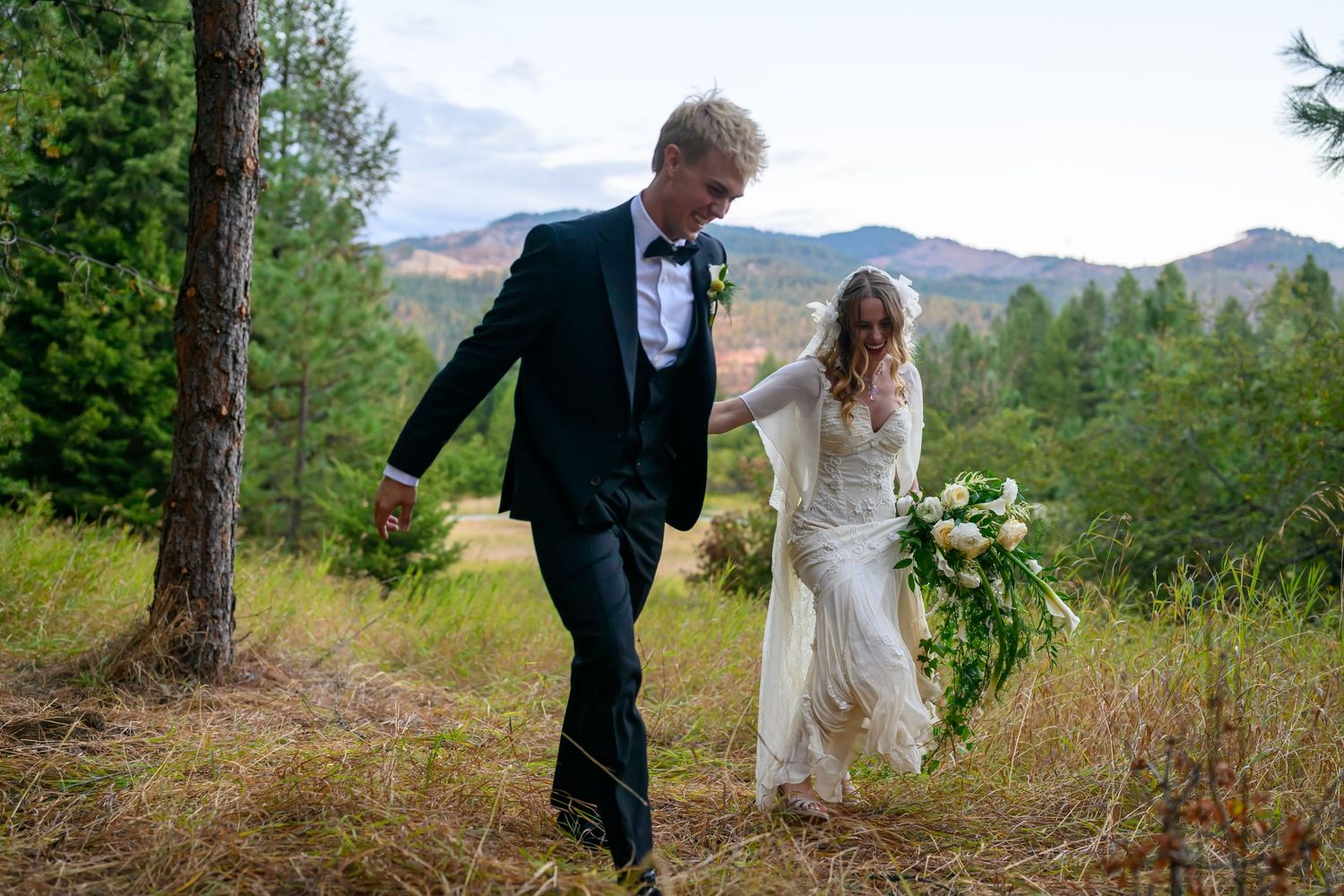 Bride and groom walking hand in hand through a mountain meadow with pine trees and peaks behind.