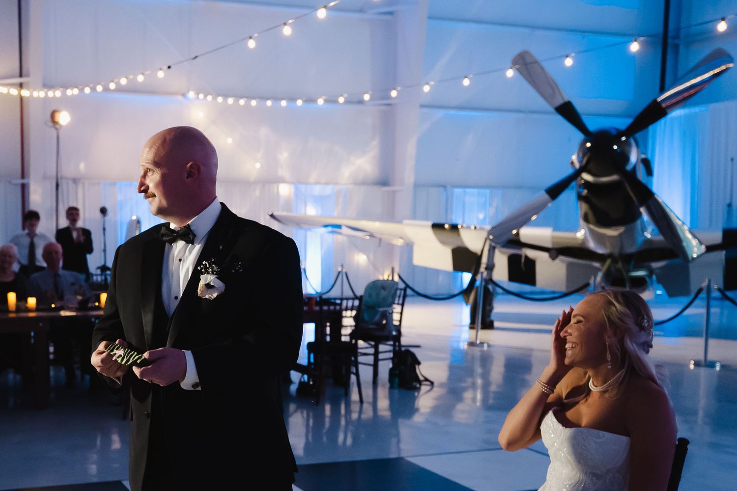 Elegant wedding reception in an aviation museum with a vintage propeller plane, string lights, and bride and groom sharing a moment.