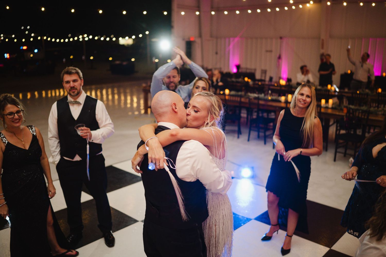 Bride and groom share a kiss on a checkered dance floor surrounded by wedding guests.