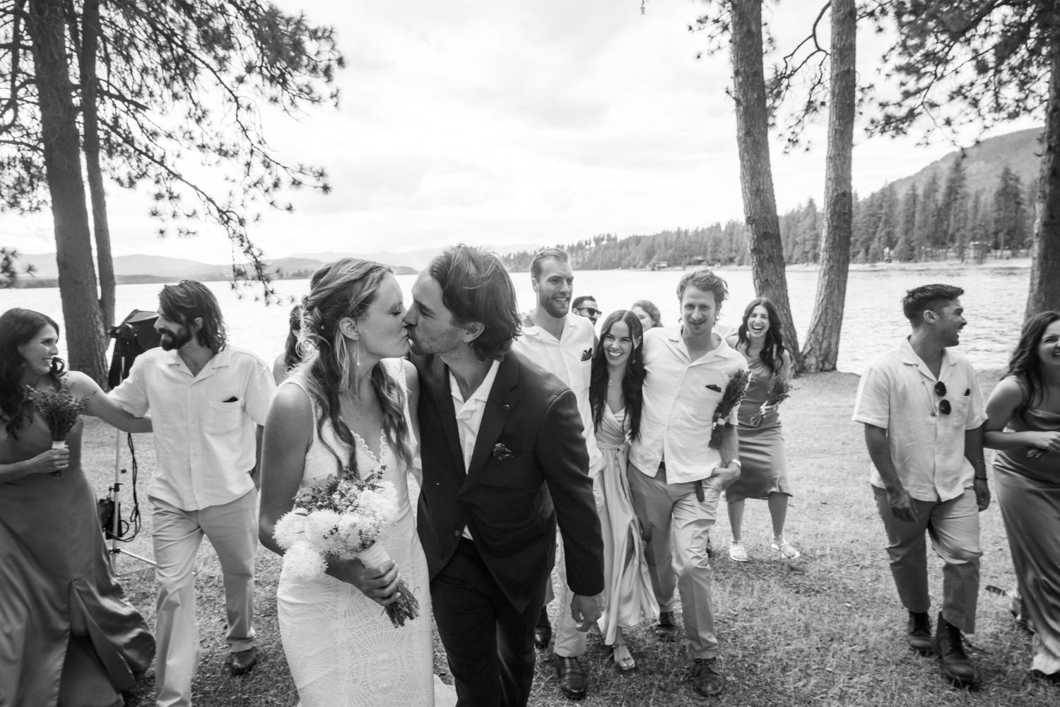 A black and white panoramic photo shows a group of people gathered outdoors by a lake for a casual celebration.
