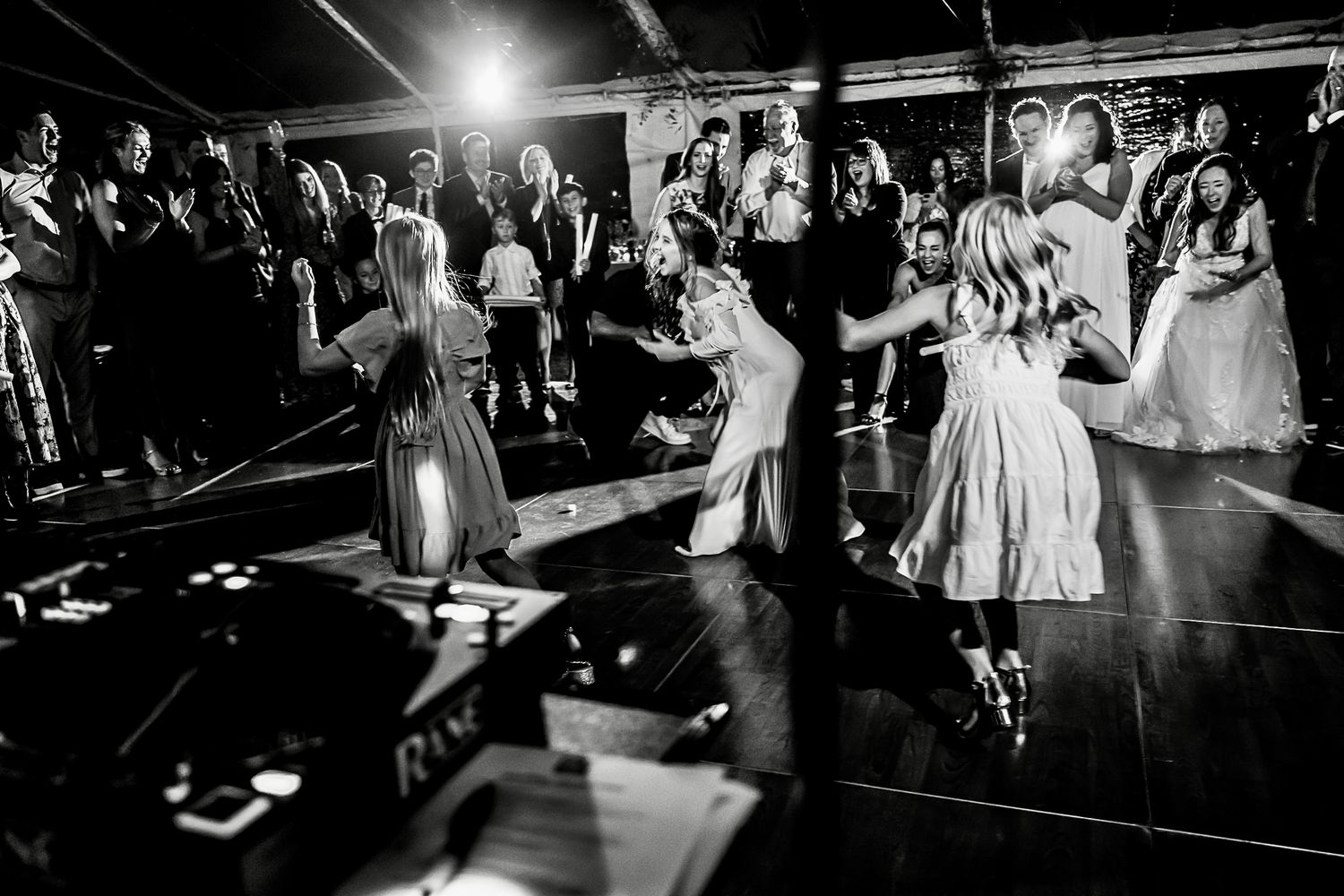 Wedding guests dance and celebrate on a dark dance floor at an evening reception in black and white photography.