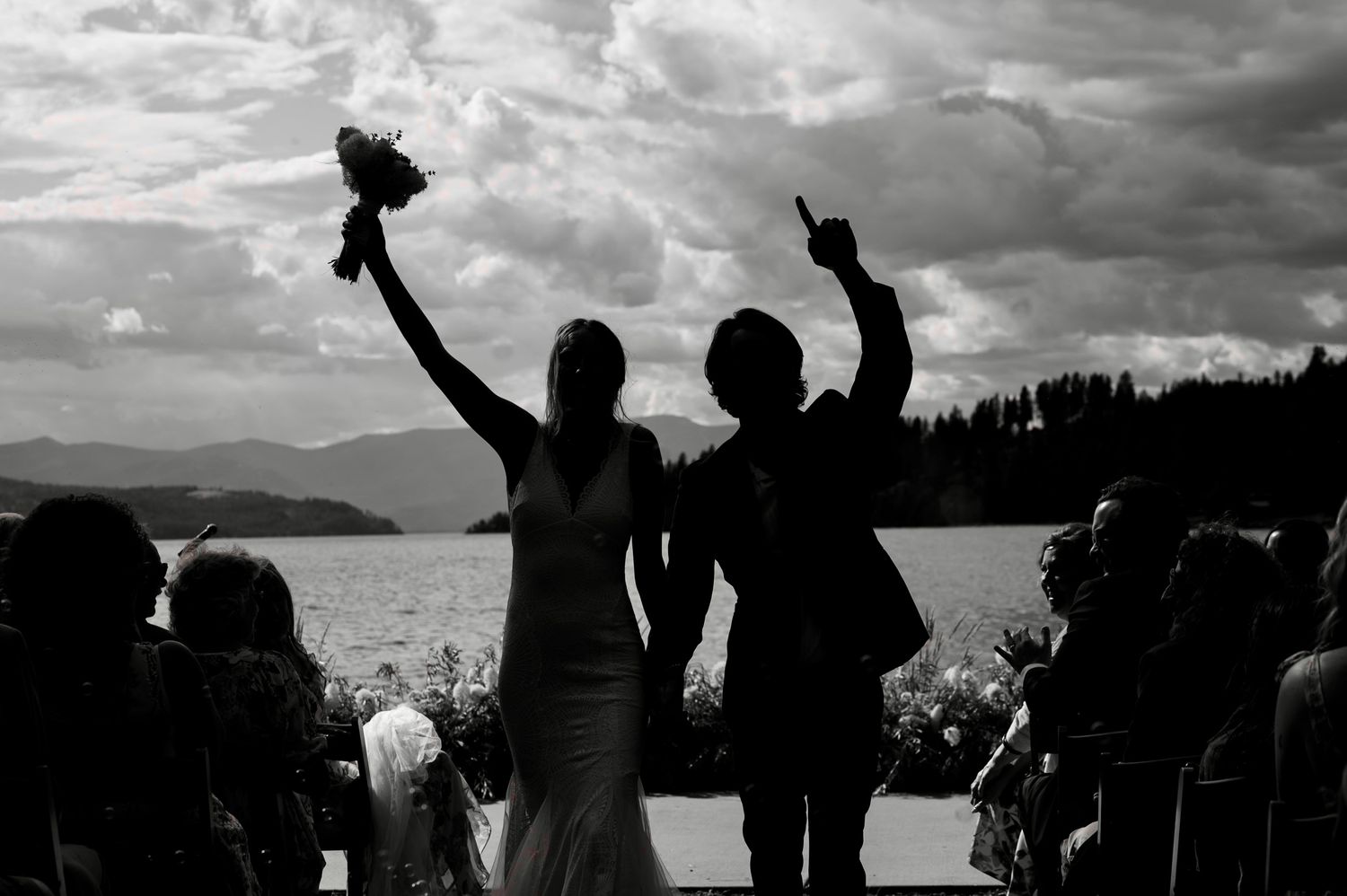 Silhouettes of a celebration by a lake with mountains in the background under dramatic cloudy skies.