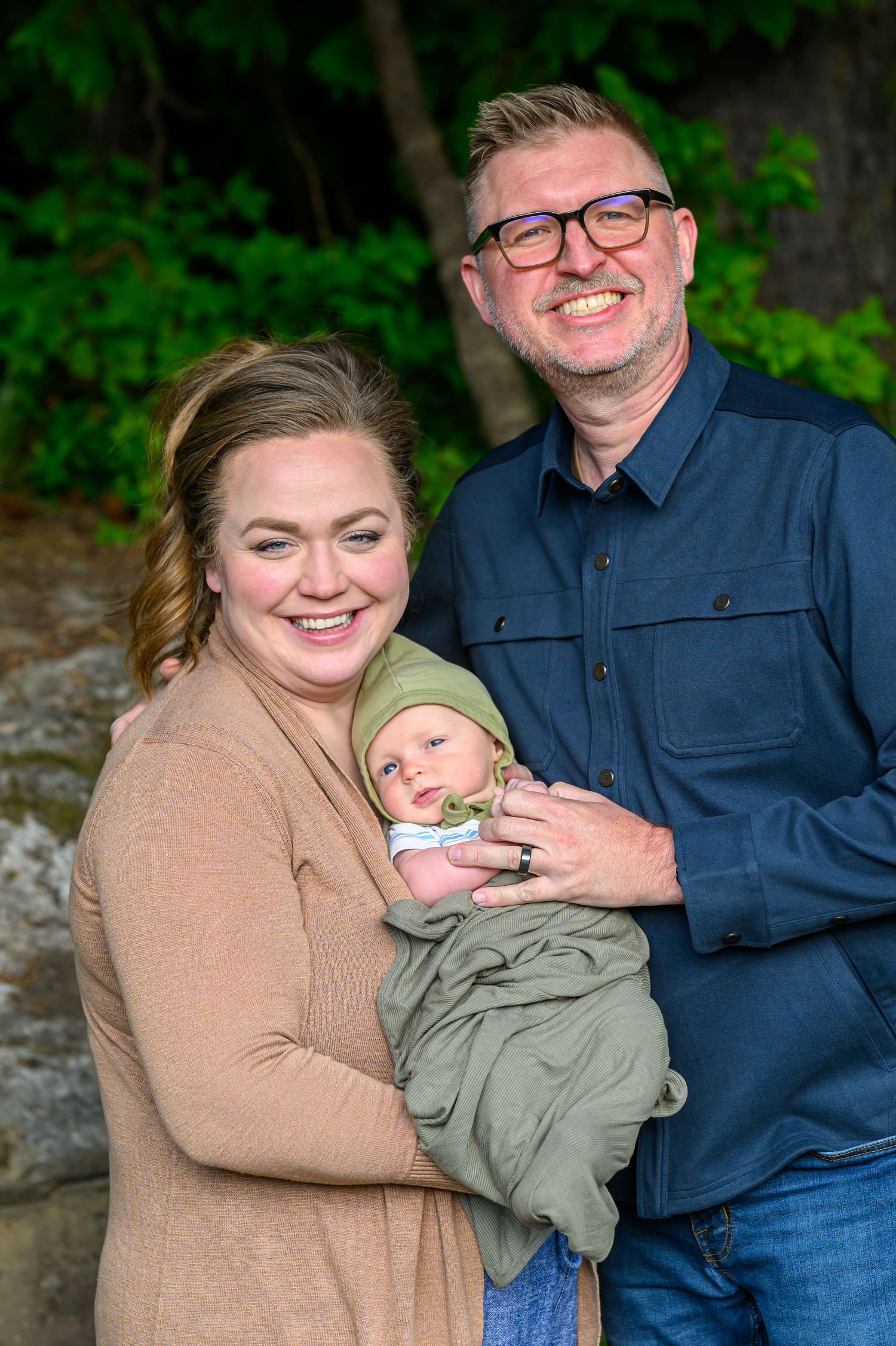 Smiling couple holds their newborn baby dressed in green outdoors among lush trees.