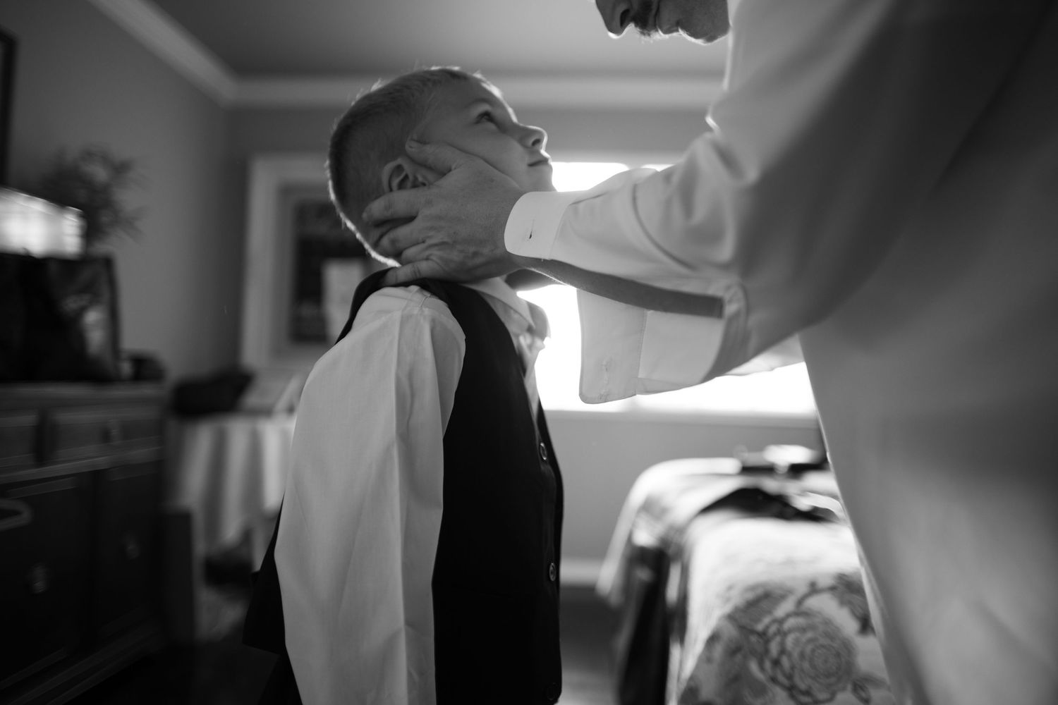 Black and white photo of a young boy having his tie adjusted by an adult during wedding preparations.