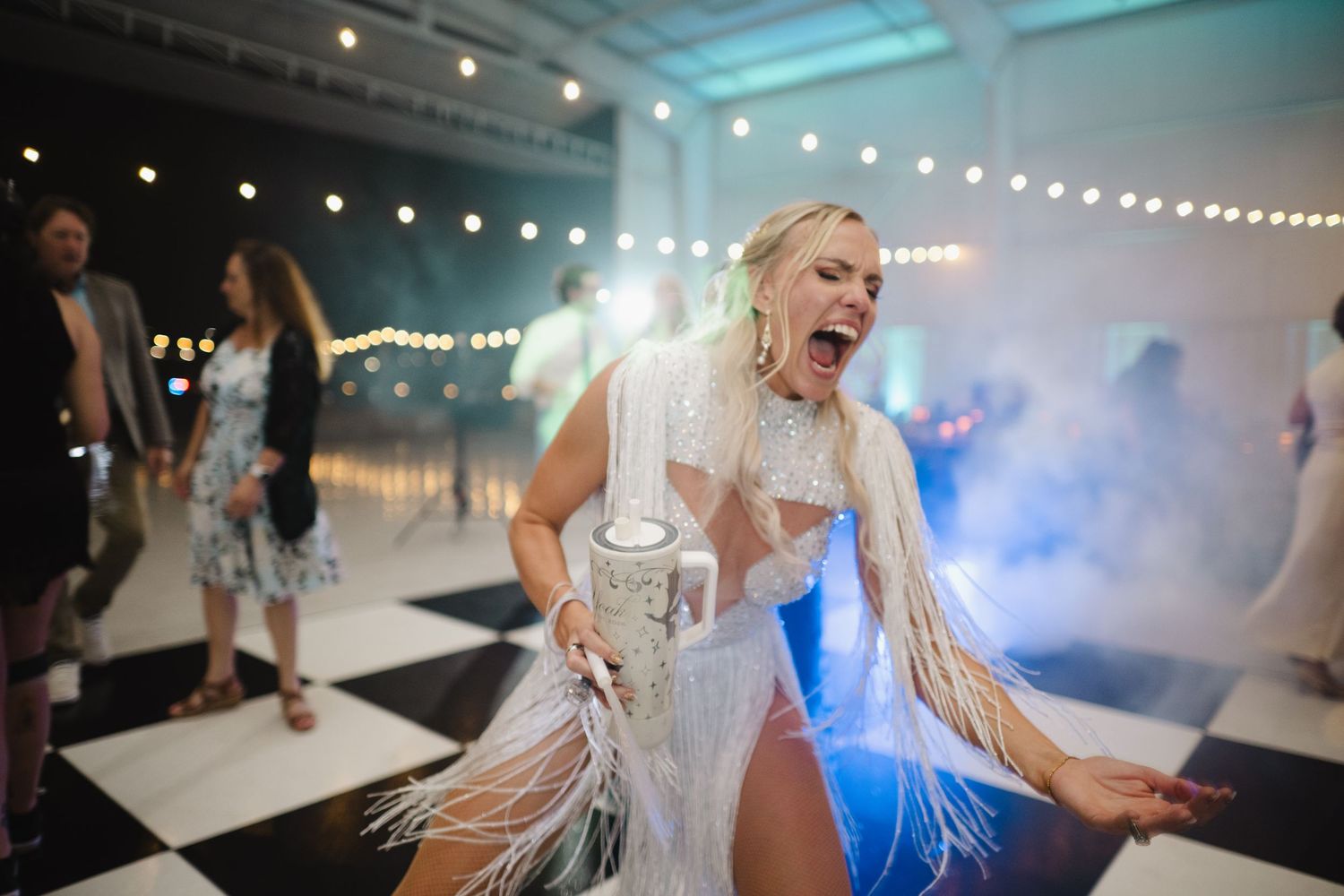 Excited blonde woman in white fringe outfit dancing on checkered floor at wedding reception.