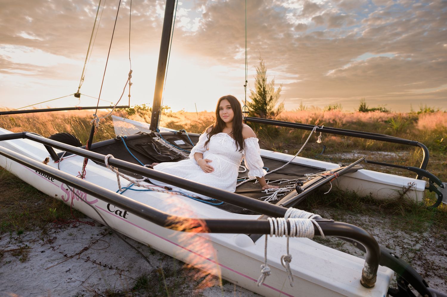 Pregnant woman in white dress poses on a catamaran sailboat at golden sunset hour on a sandy beach.