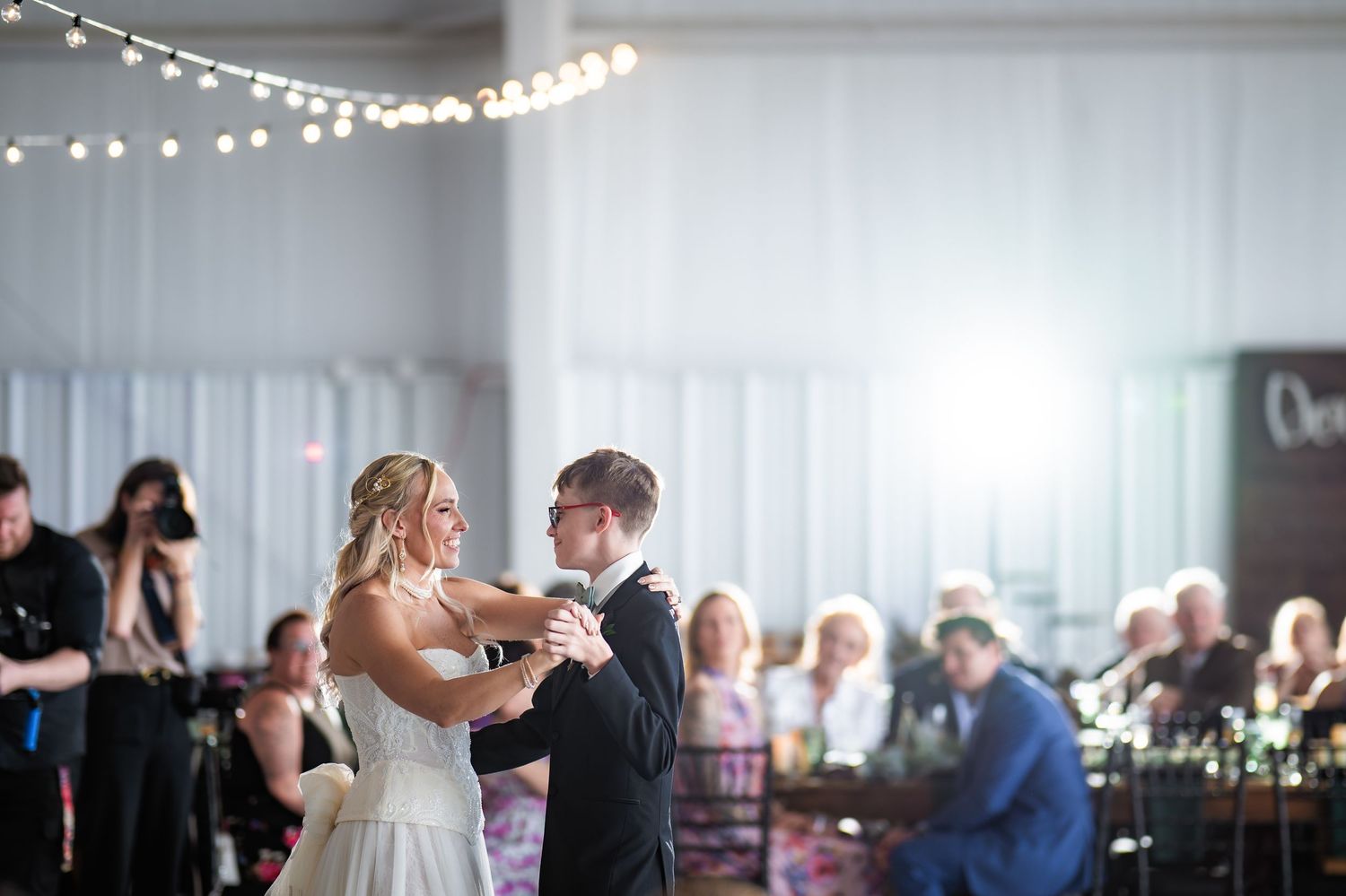 Bride and groom share their first dance at elegant wedding reception with string lights and guests seated nearby.