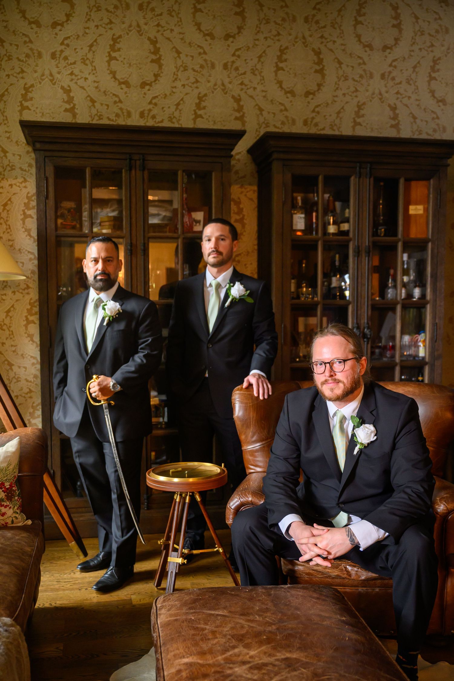 Groomsmen in black suits pose casually on vintage leather furniture in an elegant room with patterned wallpaper.