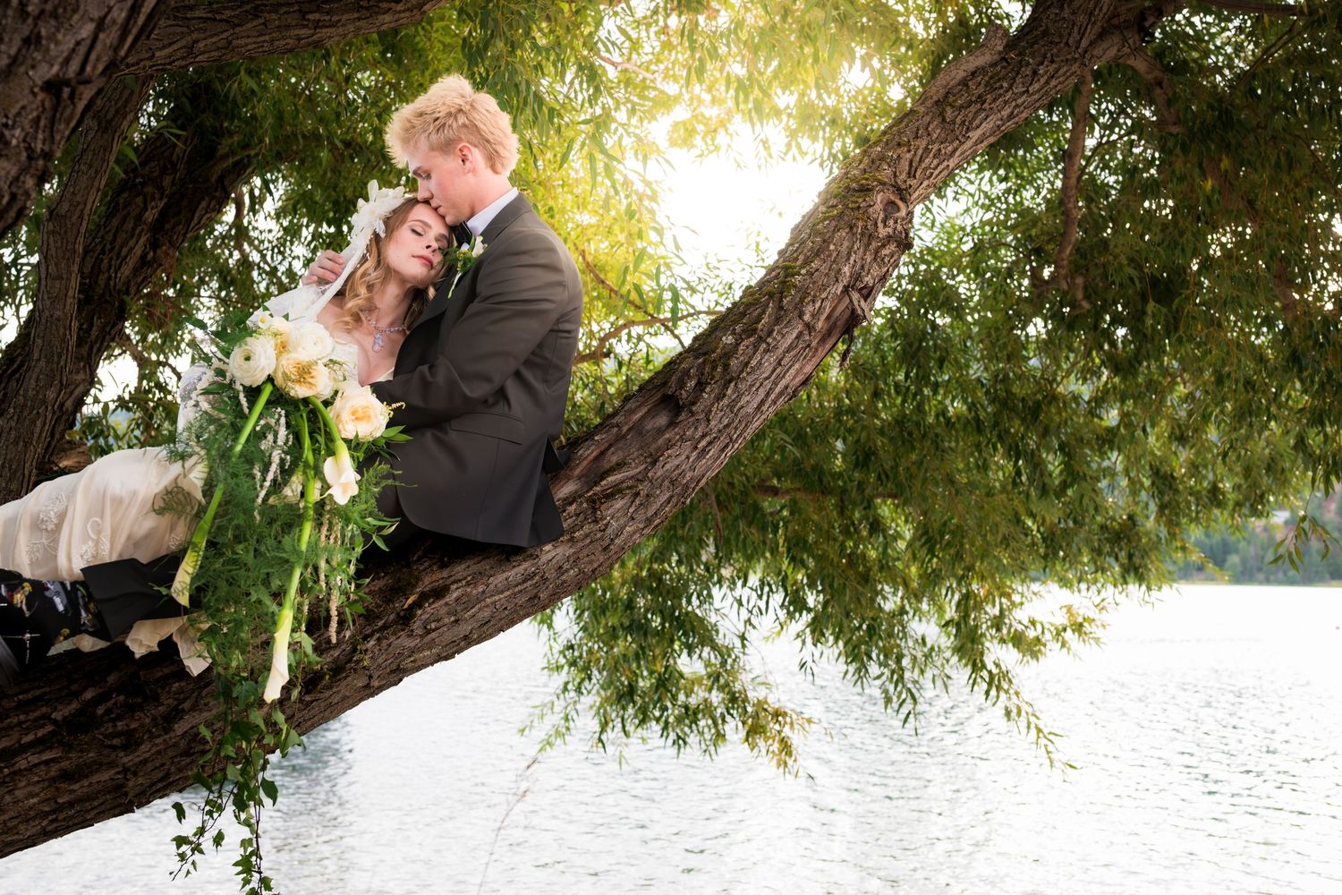 Bride and groom sitting romantically in a large tree by a lake holding a bouquet.
