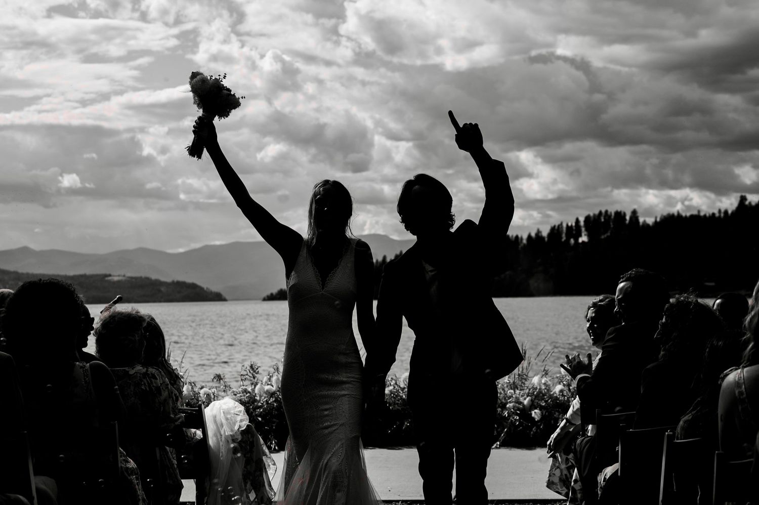 Black and white silhouette of newlyweds celebrating at outdoor lakeside wedding ceremony with mountains in background.