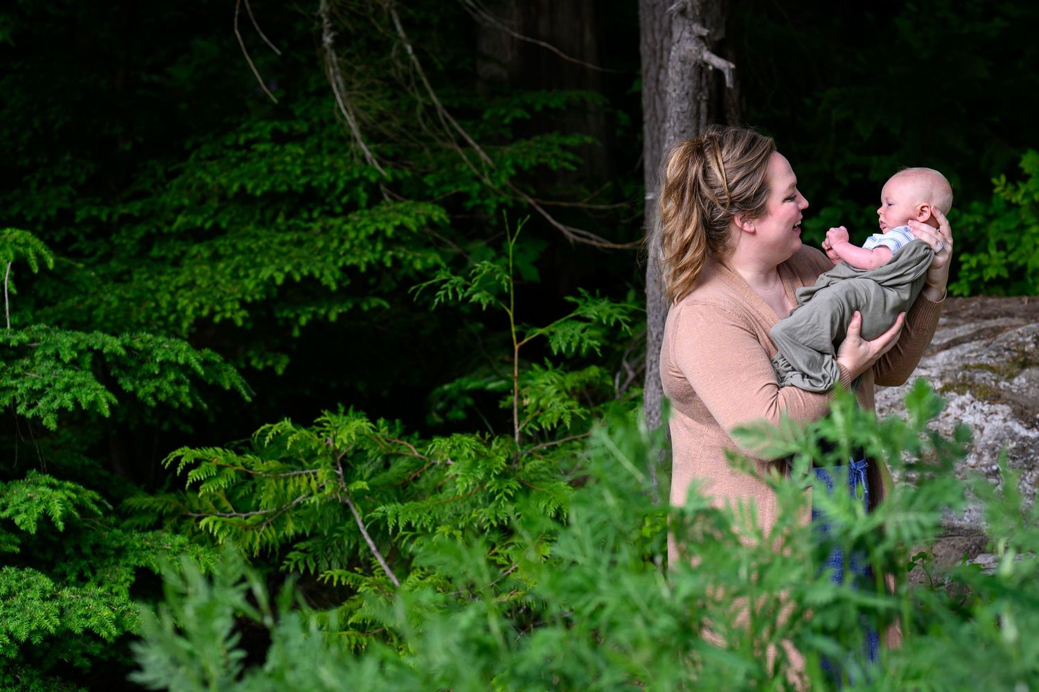 A woman holds a baby outdoors near lush green forest trees in a serene natural setting.
