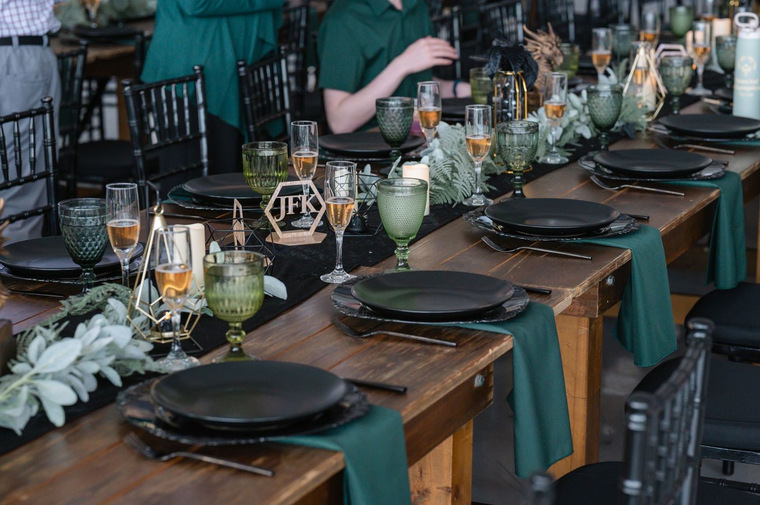 Elegant wedding reception tablescape with black plates, green runners, and geometric gold centerpieces on wood tables.