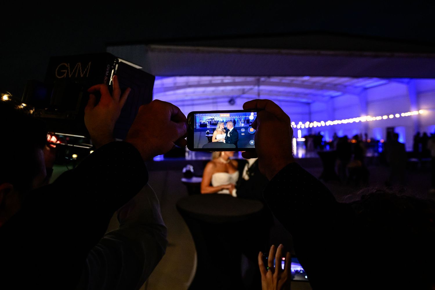 Wedding guest captures bride and groom's first dance on smartphone at outdoor evening reception.