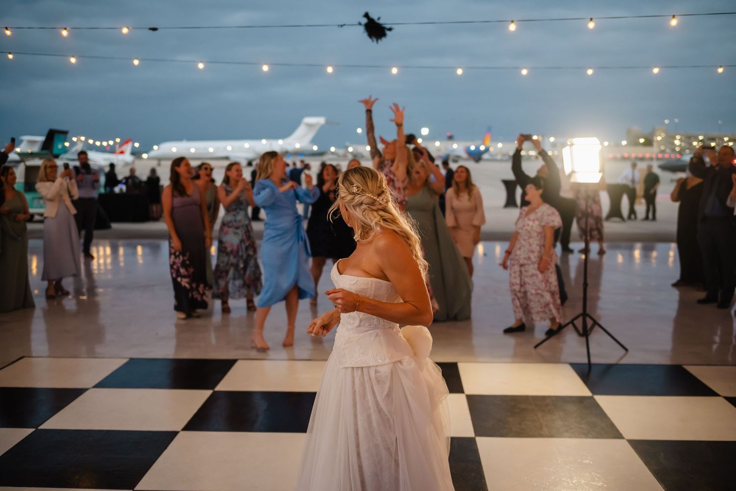 Bride tosses bouquet to guests on checkered dance floor at outdoor airport hangar wedding reception.