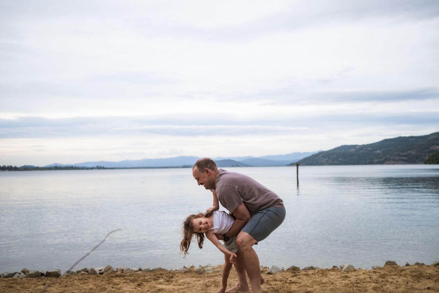 Man playfully dipping a young girl on a sandy beach with calm lake and mountains in the background.