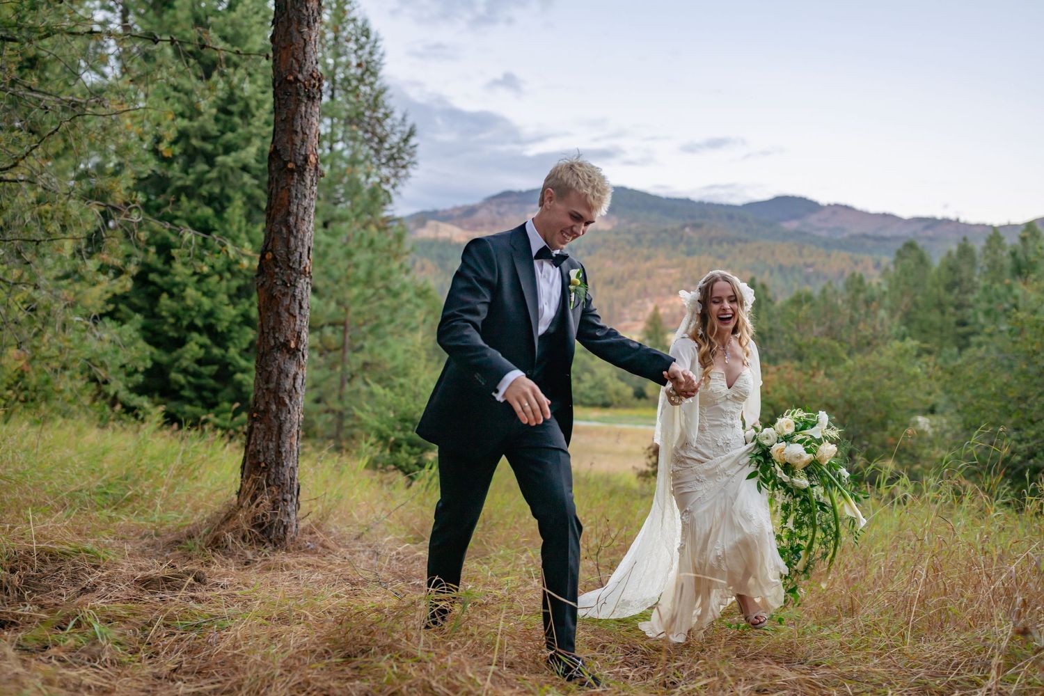 Bride and groom walking through mountain meadow, bride holding white bouquet in lace wedding dress.
