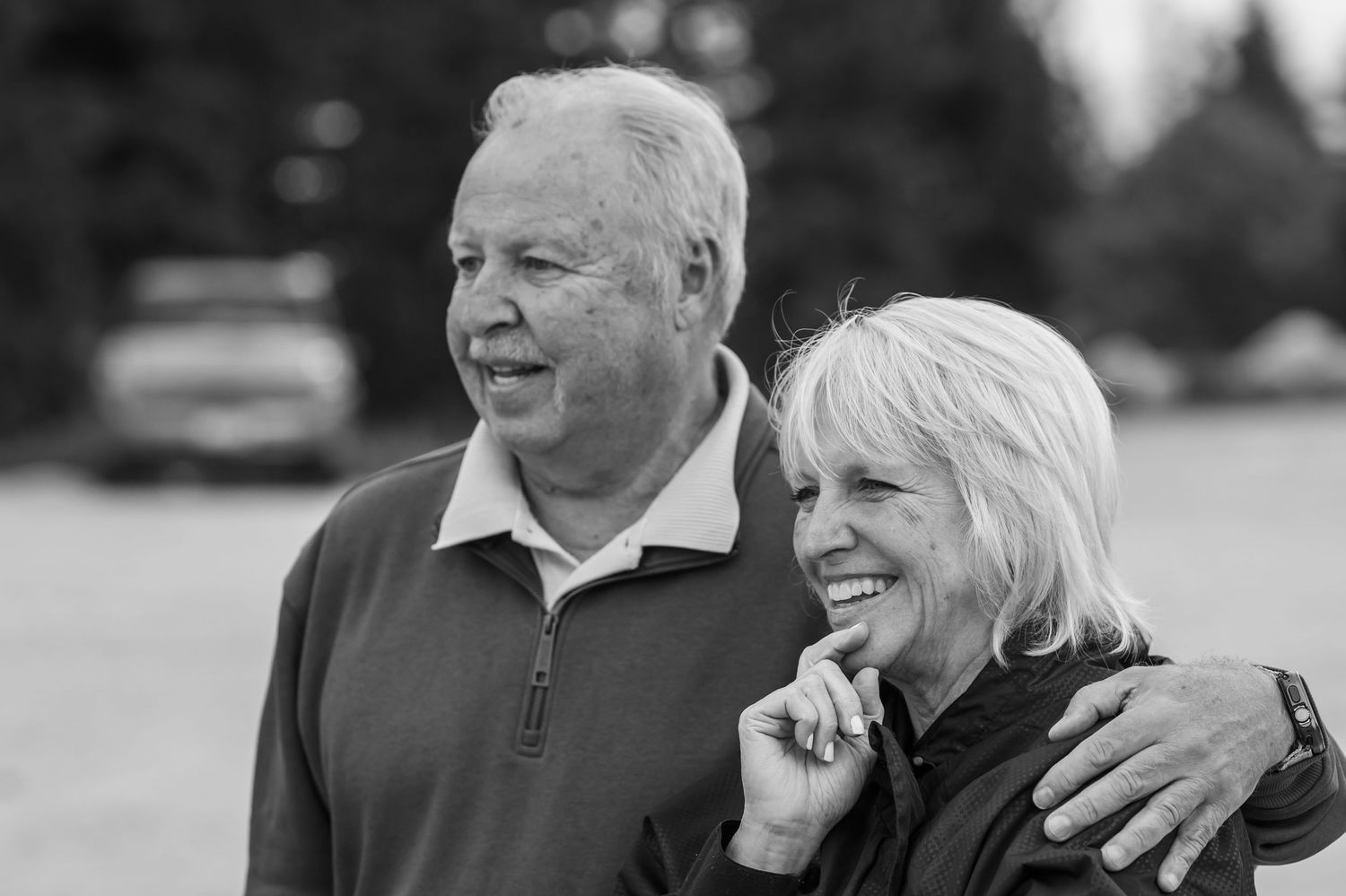 Black and white photo of a smiling senior couple outdoors, man with arm around woman, both looking cheerfully ahead.