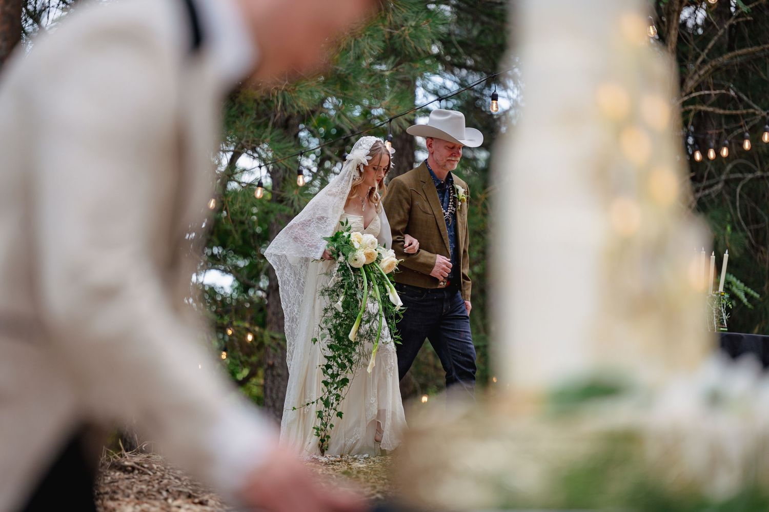 Bride in lace gown with cascading greenery bouquet walks with man in cowboy hat at outdoor wedding.