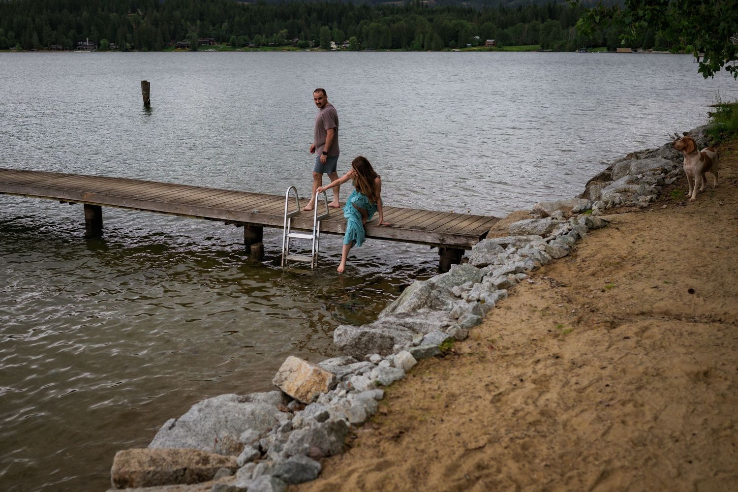 Couple walking on a wooden dock by a rocky shoreline with calm lake and trees behind.