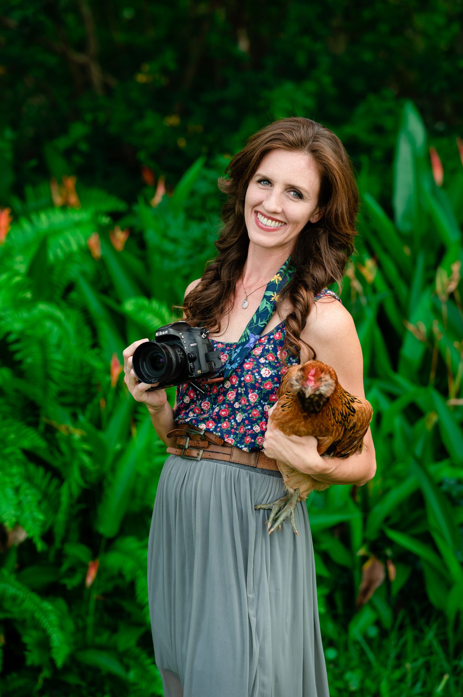 A photographer with camera equipment poses with a chicken in a lush green garden setting.