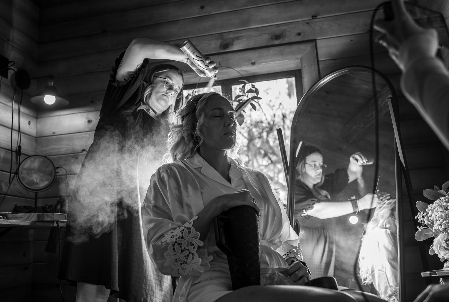 Black and white photo of a bride getting hair styled with spray in a rustic cabin before her wedding.