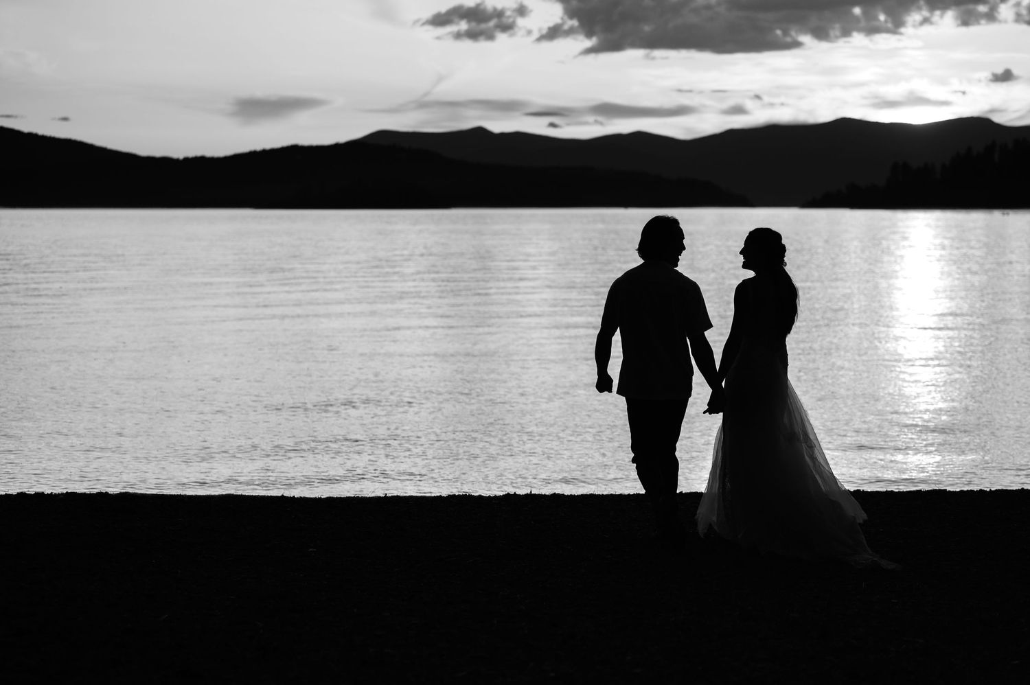 A silhouetted couple in wedding attire stand together against a serene lake backdrop with mountains at sunset.