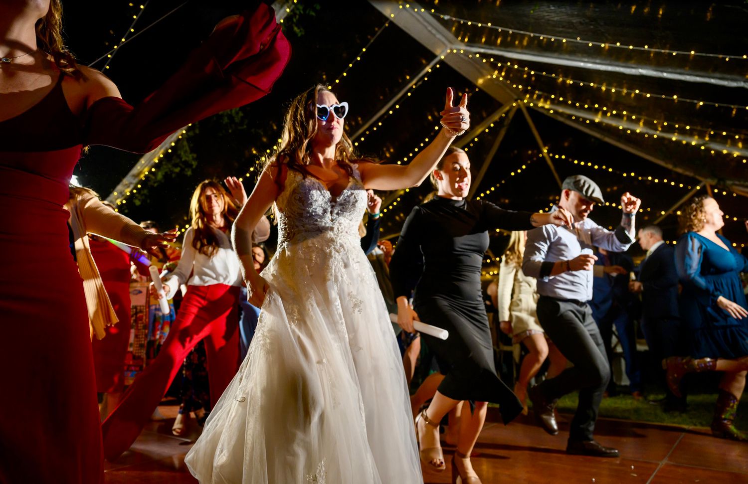 Wedding guests dance together under decorative string lights at a lively evening reception celebration.