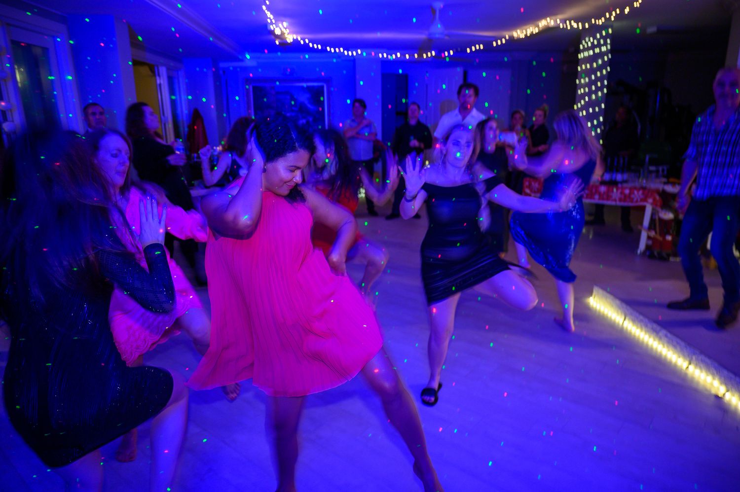 Dancers move energetically under blue lighting at a lively party with string lights overhead.