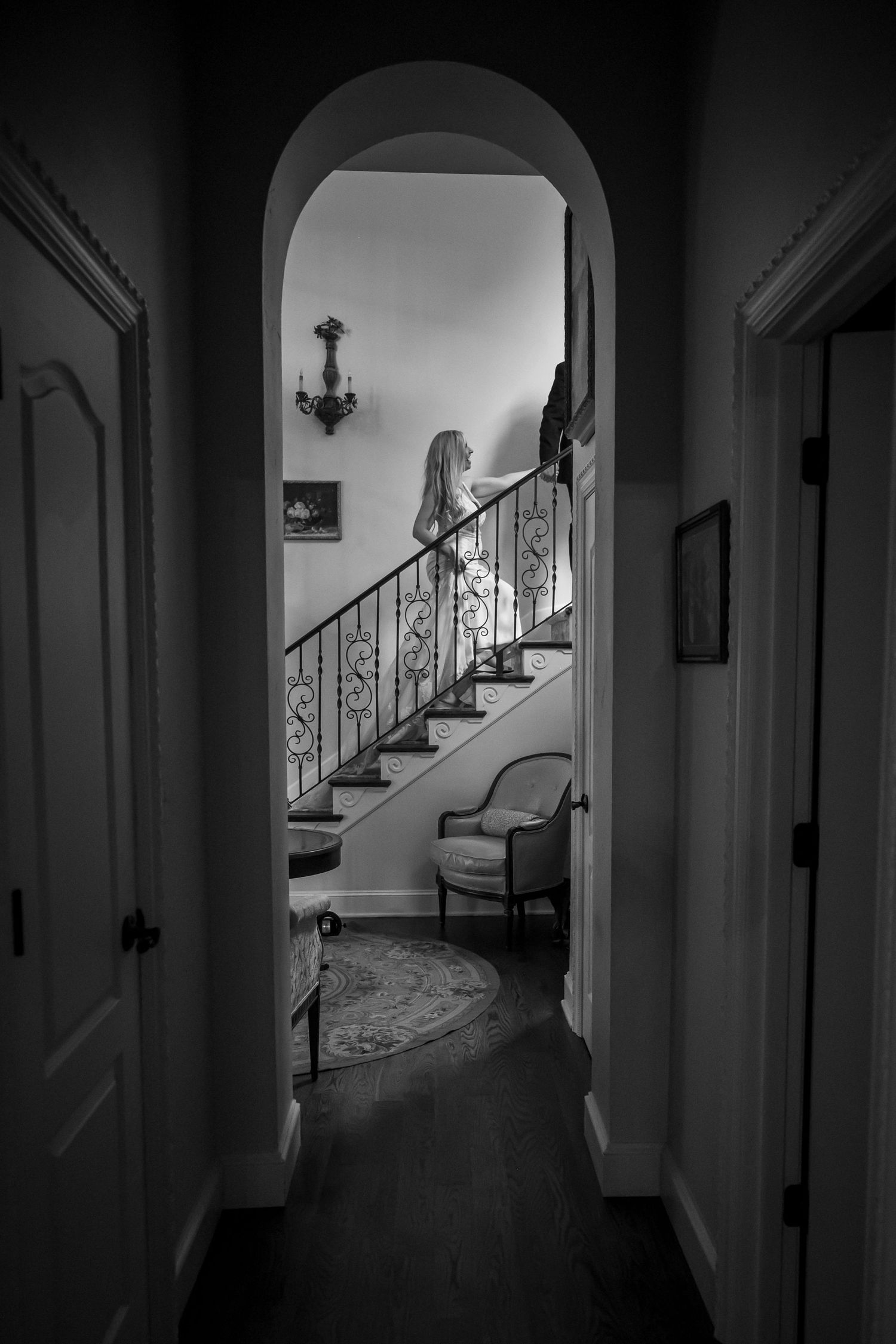 Black and white view through an arched doorway showing an elegant curved staircase with ornate railings and a wall sconce.