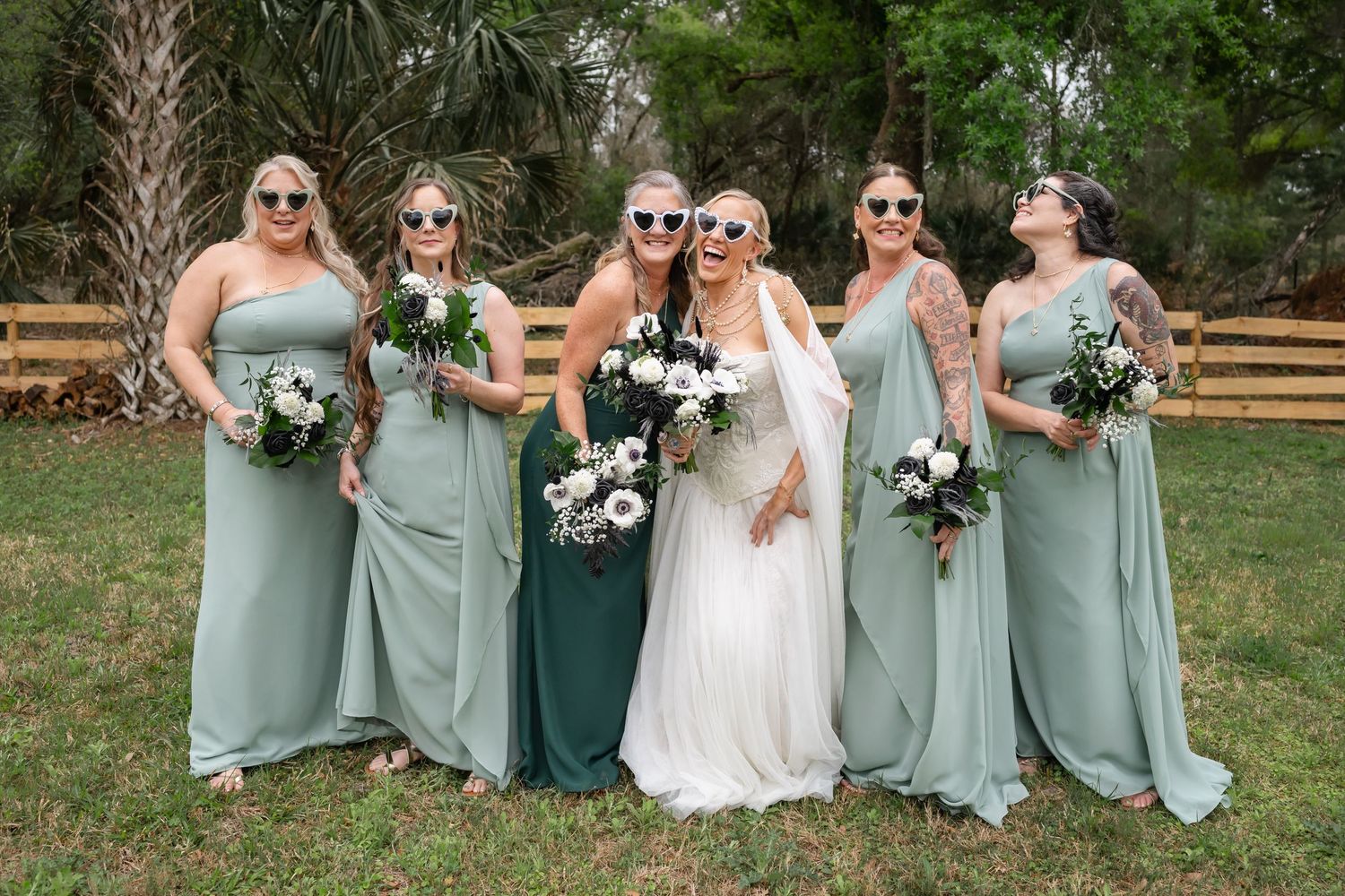Bride and bridesmaids in sage green dresses holding bouquets of white and green flowers outdoors.