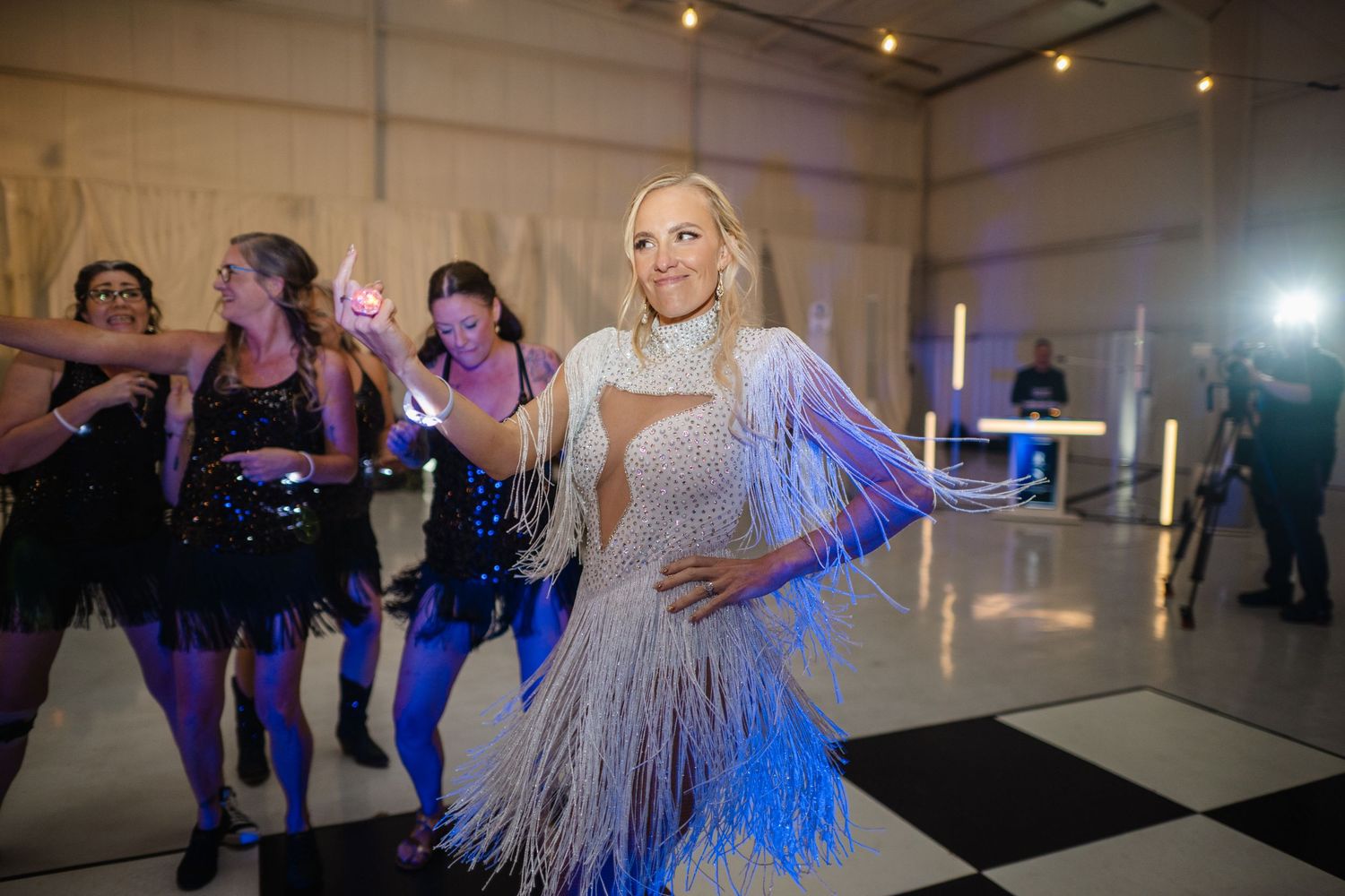 Woman in white fringed dress dancing at elegant party with performers in black sequin outfits on checkered dance floor.