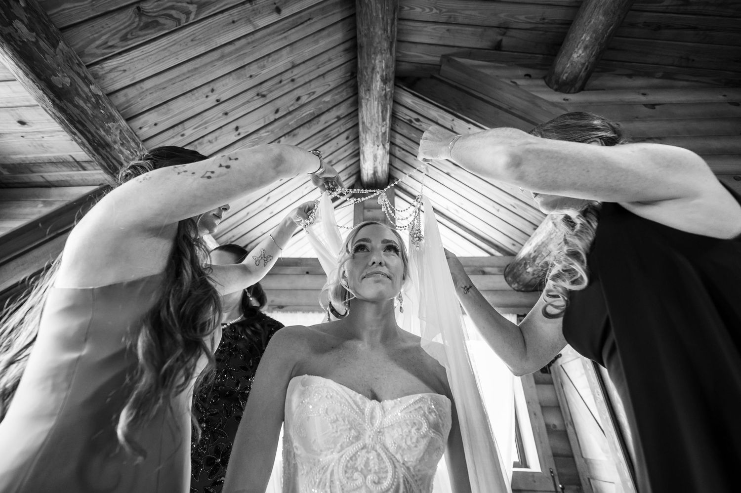 Black and white photo of bridesmaids placing veil on bride in rustic wooden venue.