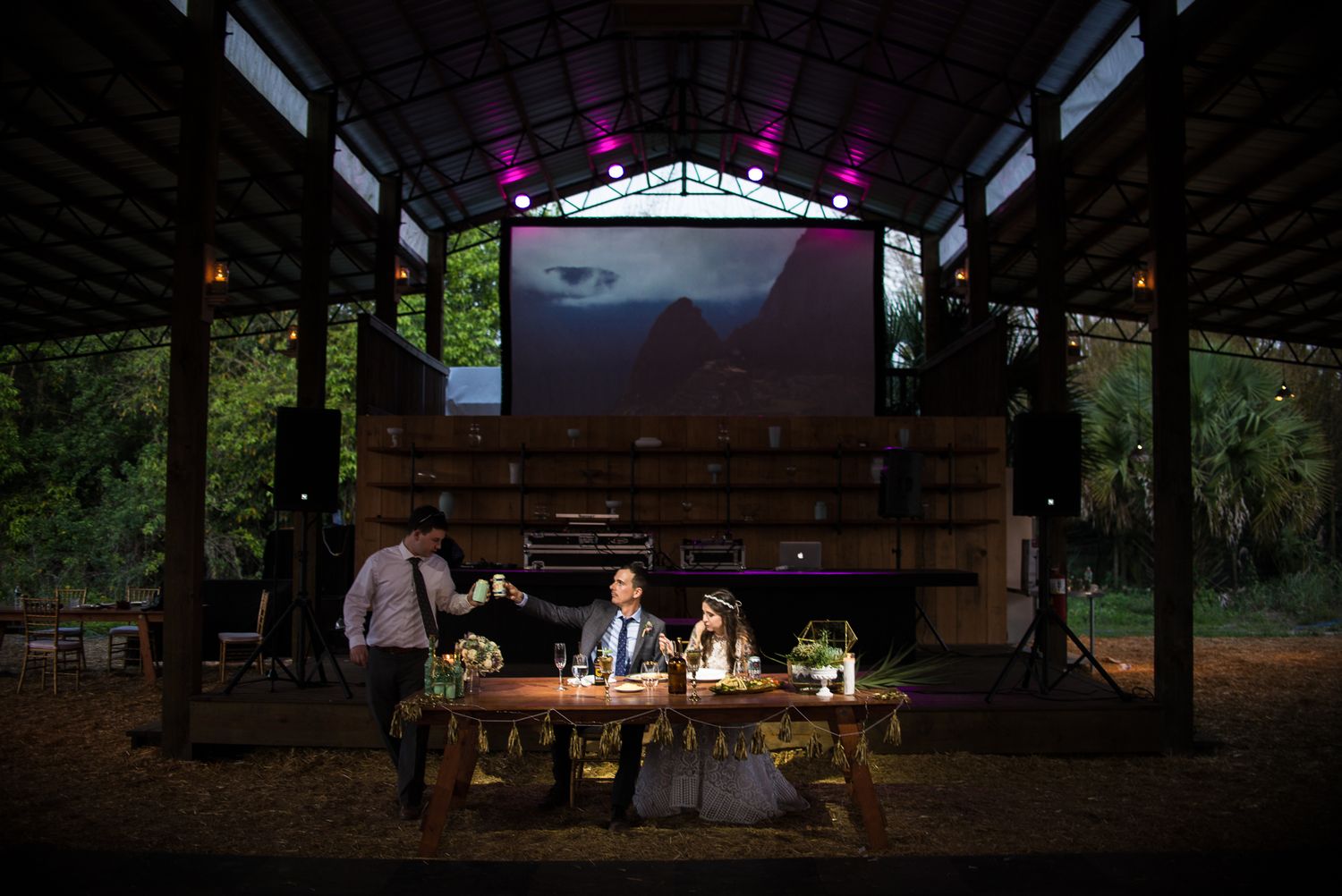 Outdoor wedding reception at dusk with glowing string lights illuminating tables under a rustic wooden pavilion.