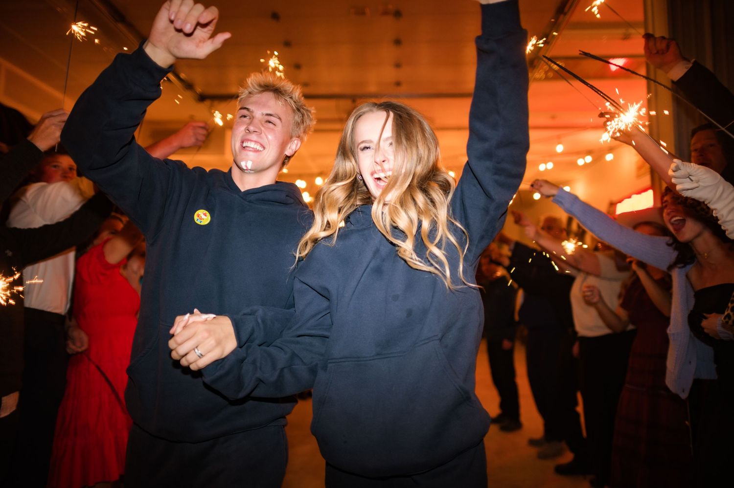 Newlyweds in matching navy sweatshirts celebrate with sparklers surrounded by wedding guests indoors.