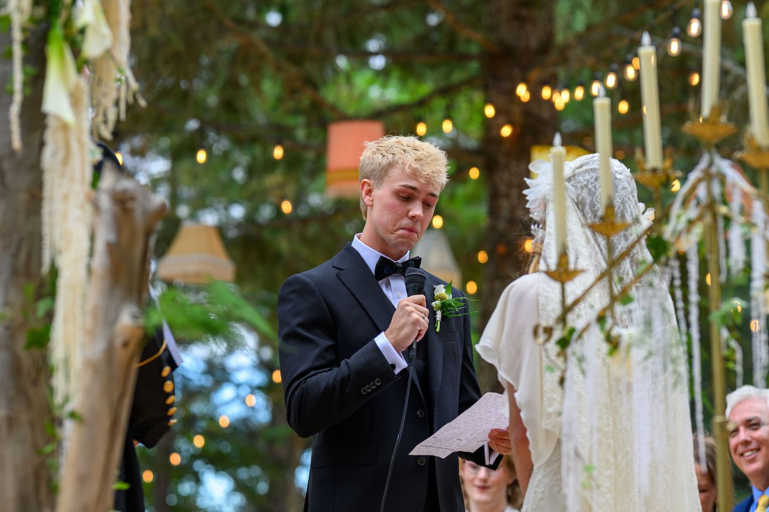 Young man in tuxedo reading vows at outdoor wedding ceremony with string lights and floral arch.