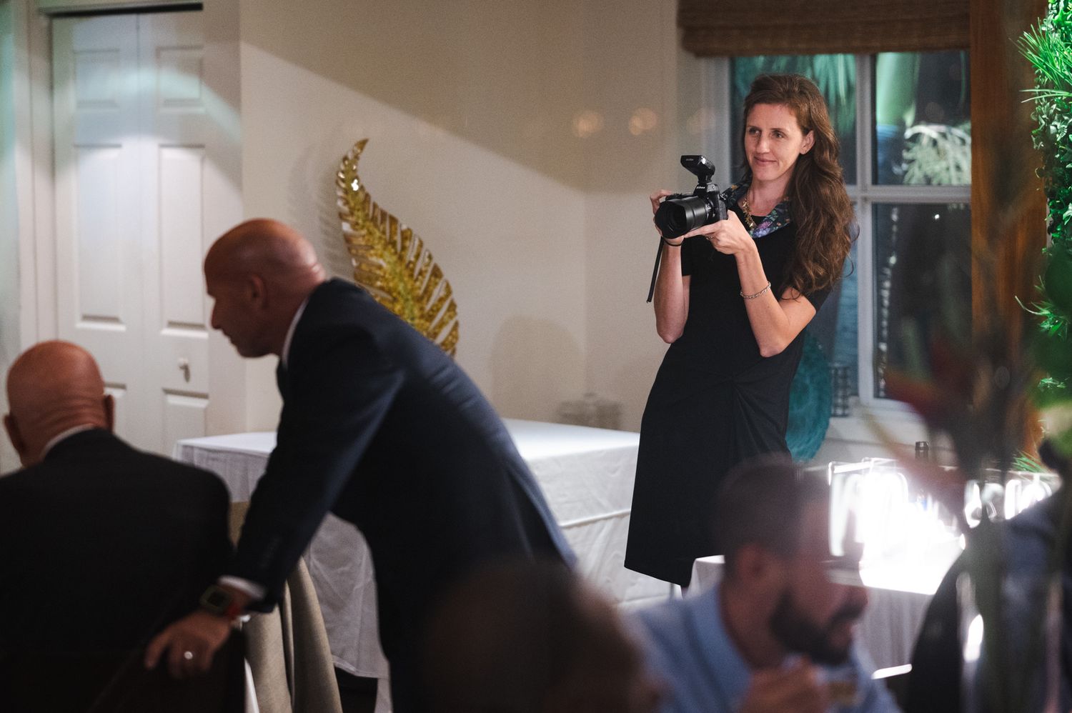 A photographer in a black dress captures candid moments at an indoor event venue with guests seated at tables.
