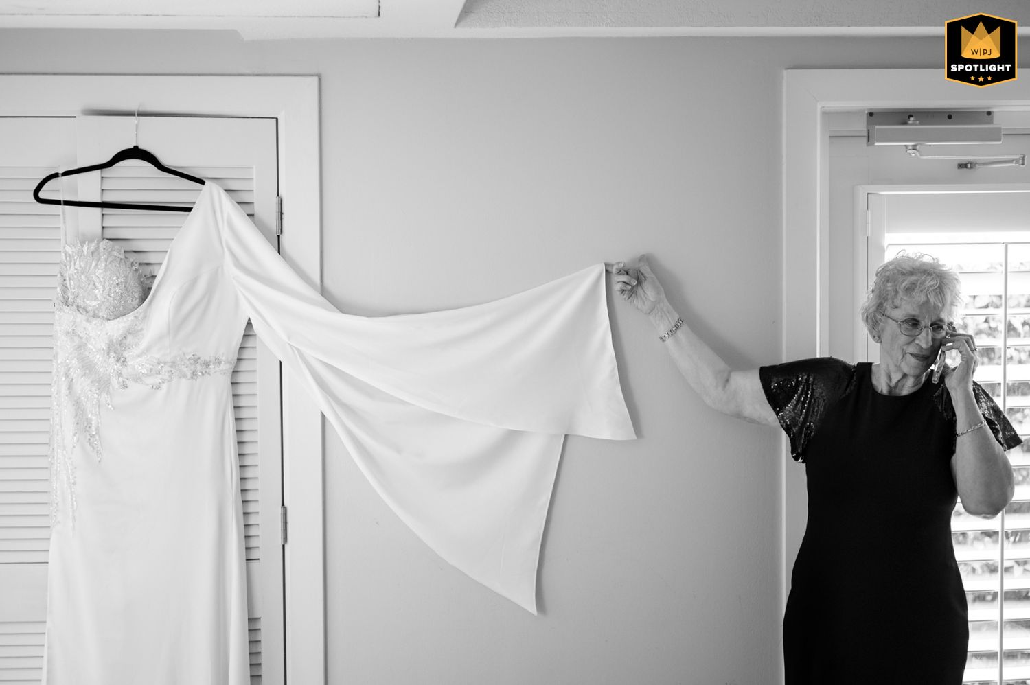 A black and white photo shows someone holding up a flowing white dress or garment against a light wall.