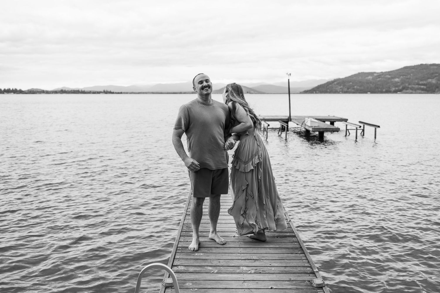 Black and white photo of couple standing and smiling together on a wooden lake dock.