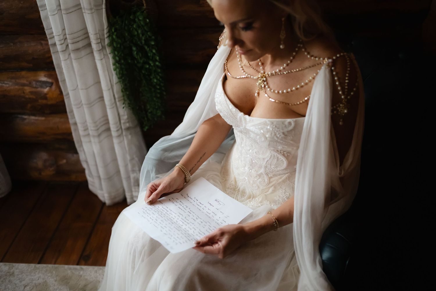 Bride in lace wedding dress and pearl necklace reads a heartfelt letter while seated in a rustic log cabin.