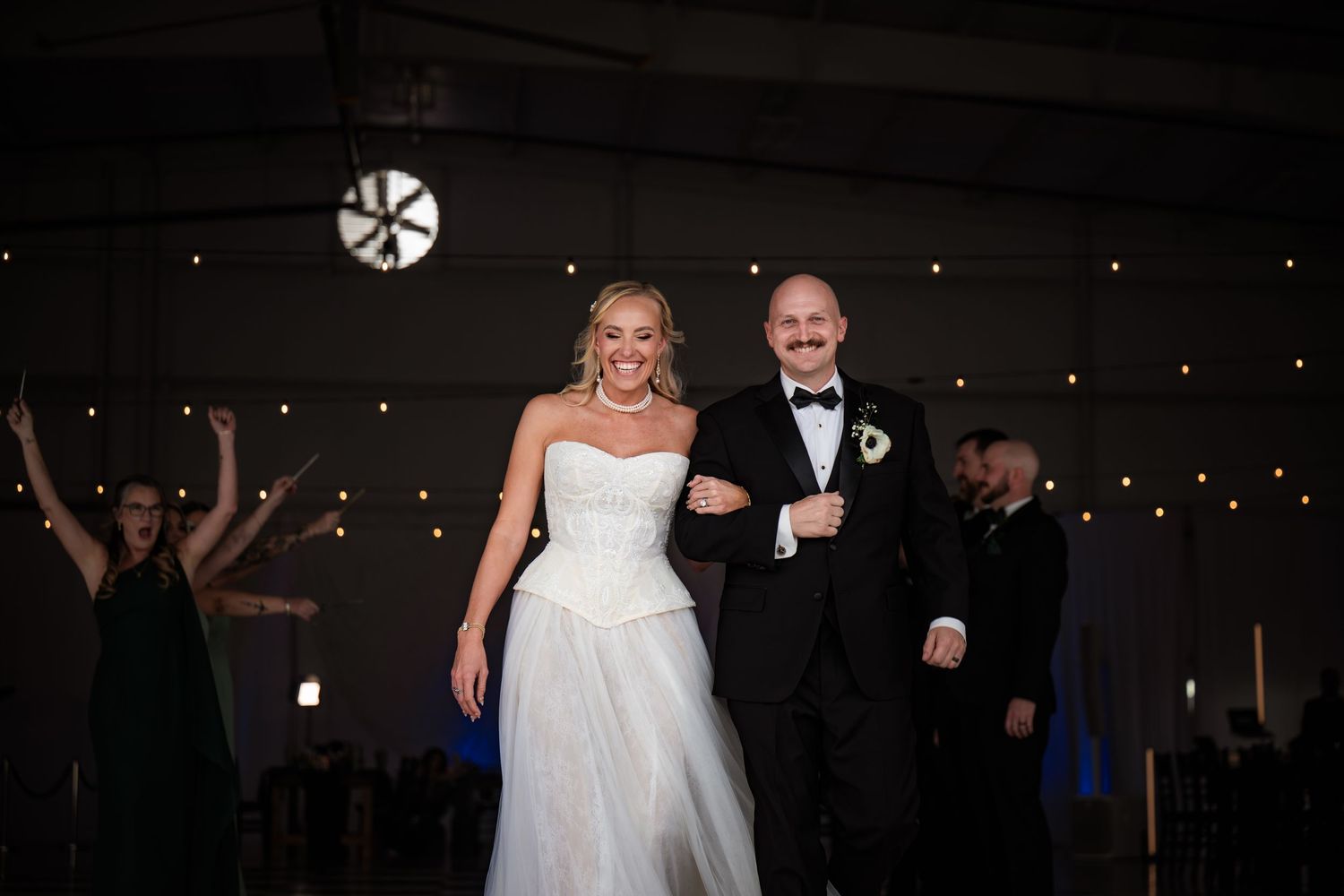 Bride in strapless white gown and groom in black tuxedo smile during wedding reception entrance under string lights.