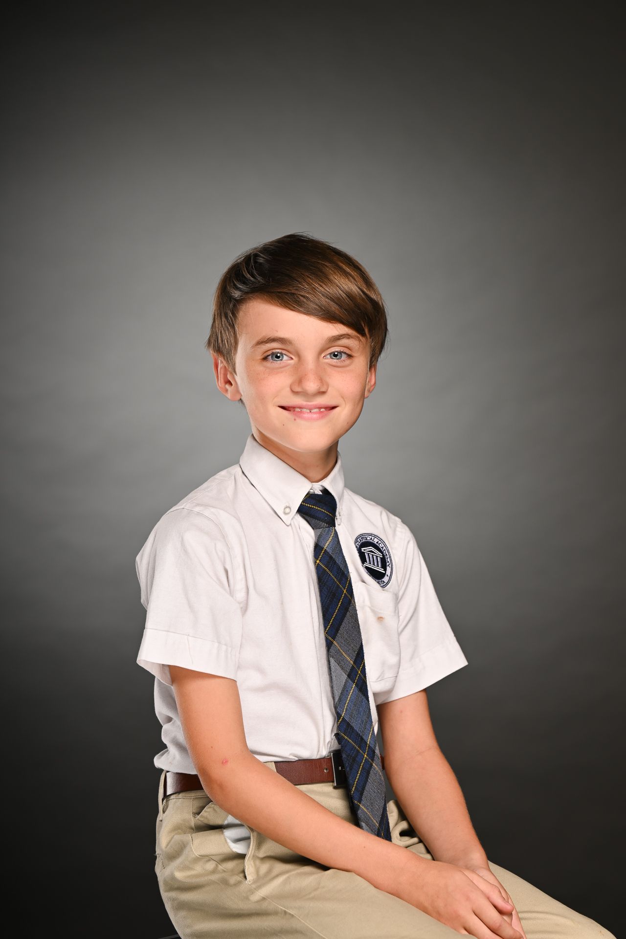 Smiling boy in school uniform with white shirt, plaid tie, and khaki pants posed for a portrait photo.