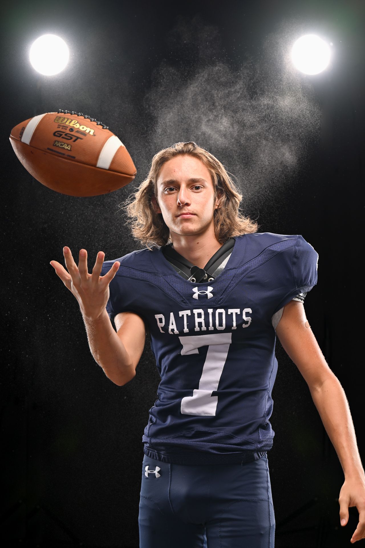 A Patriots #7 football player with long hair tosses a Wilson football under dramatic stadium lights against a dark background.