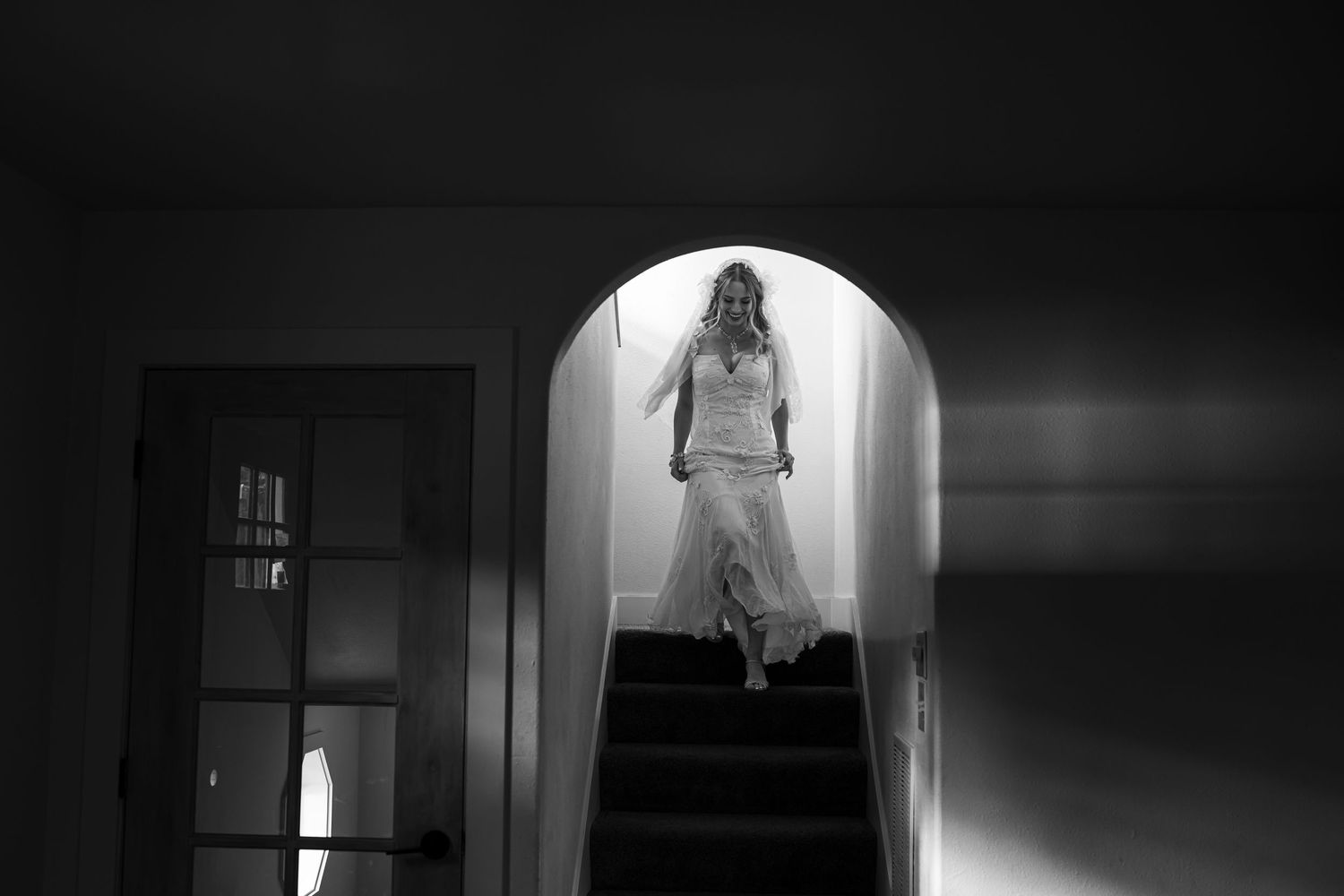 Black and white photo of a bride in a wedding dress descending a staircase through an arched doorway.