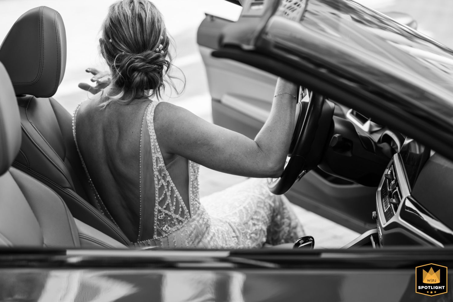 A black and white photo shows bride in a lace wedding dress sitting behind the steering wheel of a luxury convertible car with updo hair