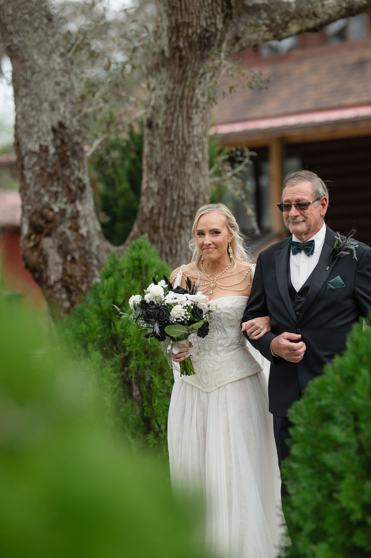Bride in white gown holding black and white bouquet walks with father during outdoor wedding ceremony.