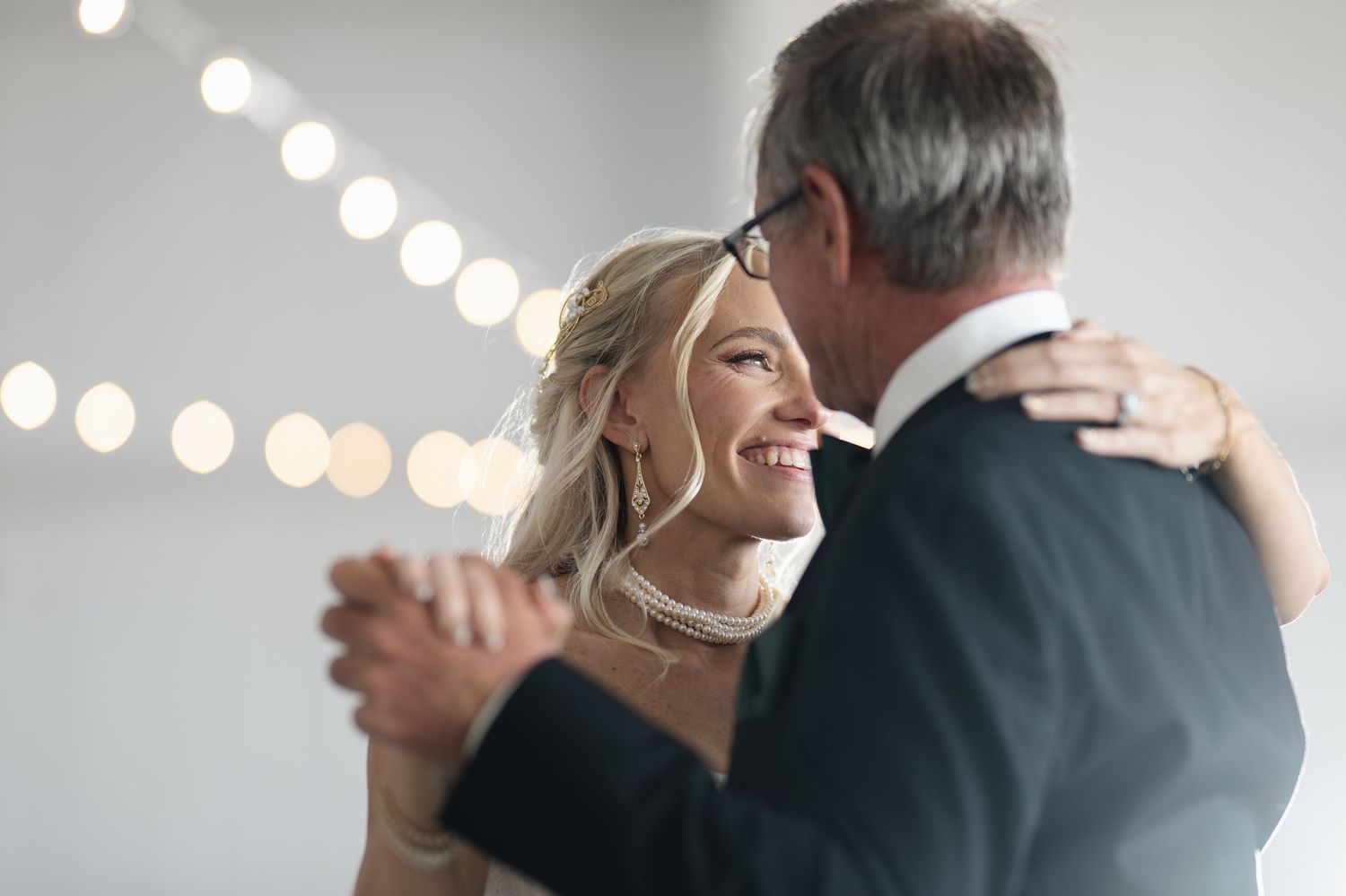 Smiling bride dances with older gentleman under warm string lights at wedding reception.
