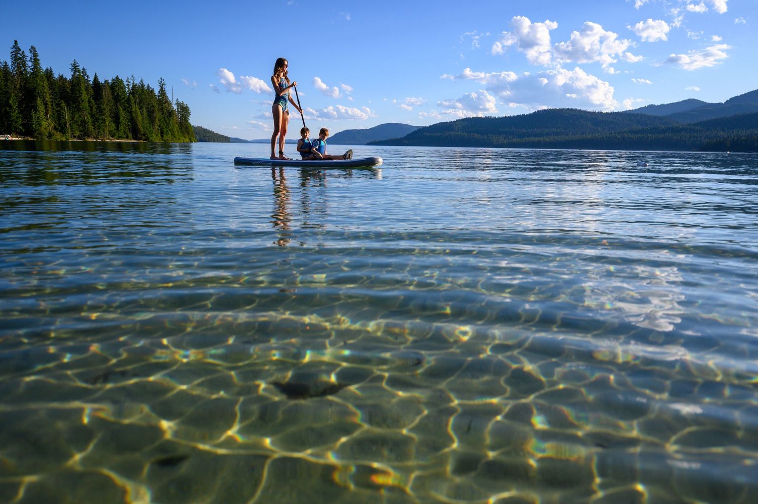 A tranquil mountain lake with clear water and evergreen trees reflects the blue summer sky in Idaho.