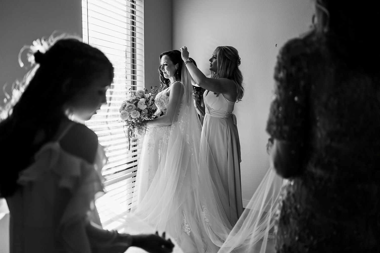 Wedding party members assist with adjusting a flowing white bridal veil near a window.