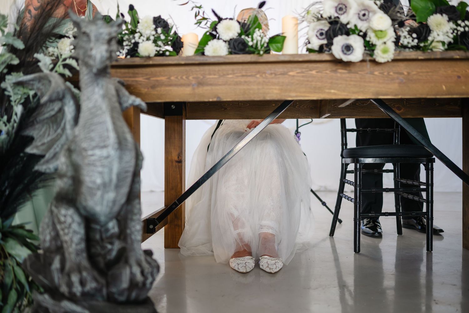 Bride in white gown sitting under rustic wood table with floral arrangements and dragon statue at fantasy wedding.